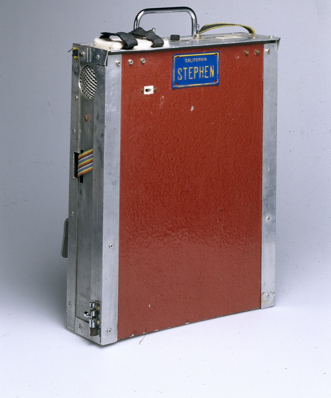 A metal-edged case with a red textured front panel and a carrying handle on top, holding the electronics. A blue California vanity license plate reading "STEPHEN" is mounted on the front, above a small rectangular window and a ribbon cable running out the side.
